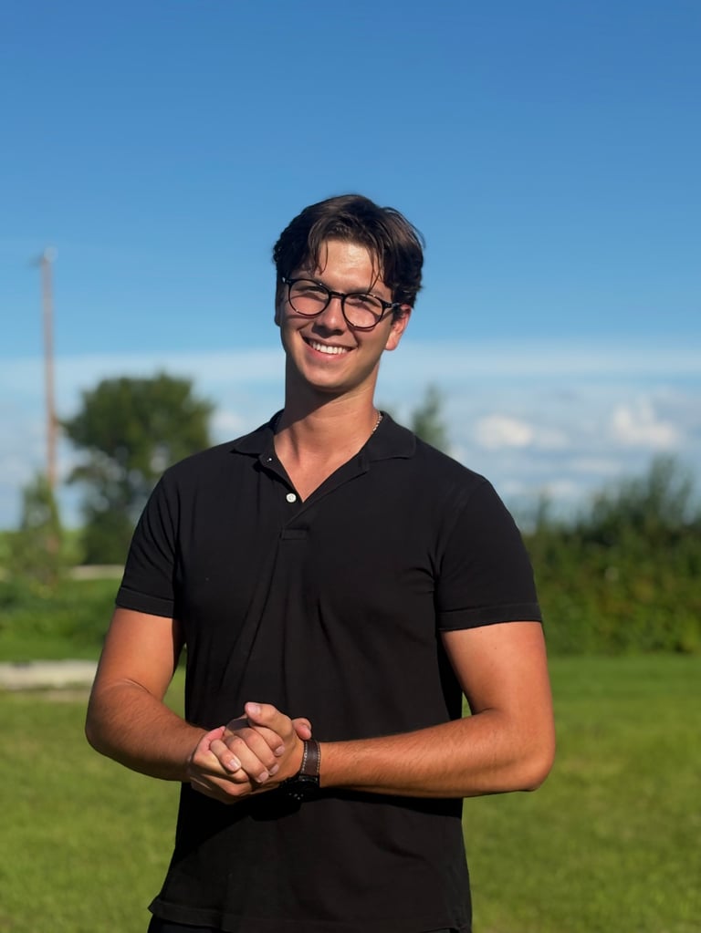 Young man wearing glasses and black v-neck shirt standing outdoors with green grass and trees in background